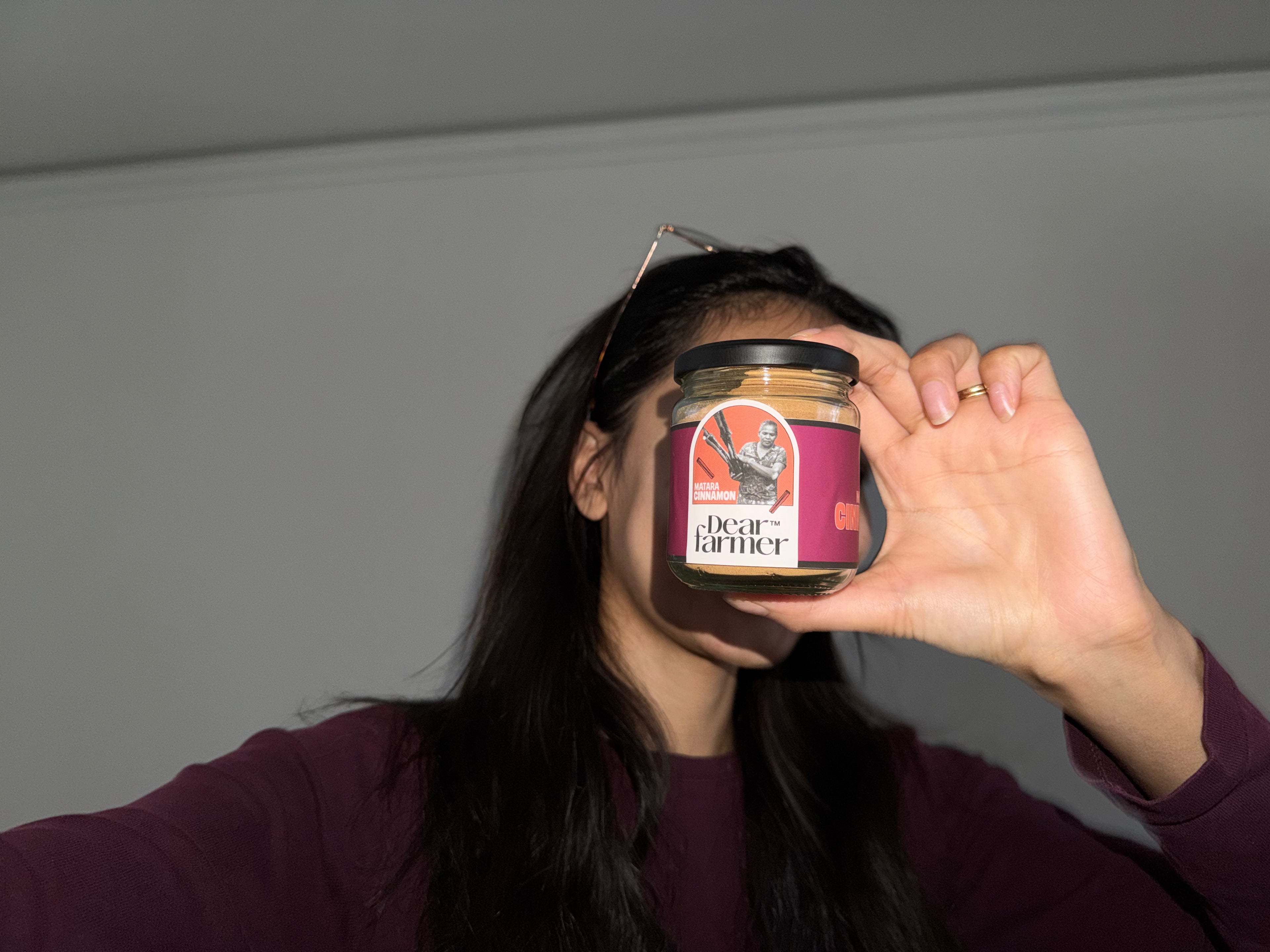 Person holding a jar of matara cinnamon by dear farmer in front of their face against a plain background